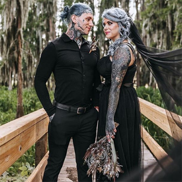 A bride with many tattoos and a gorgeous silver braid stands on a wooden bridge with her groom while holding her bouquet down at her waist. Her black veil billows out toward the camera.