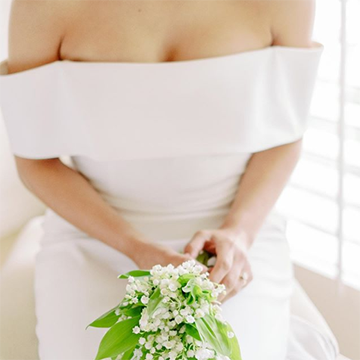 A bride in a flat, off-the-shoulder wedding dress holds a bouquet of white lily of the valley in her lap while sitting in front of a white window.