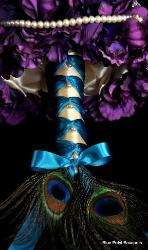 A close-up of the underside of a bouquet made of purple flowers, with a blue and white ribbon handle that has peacock feathers poking out of the butt of the handle.