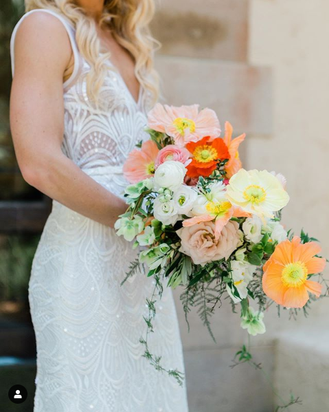 A bride holds a bouquet of light peach, white, orange, pink, and red poppies with various greenery in front of her white dress.