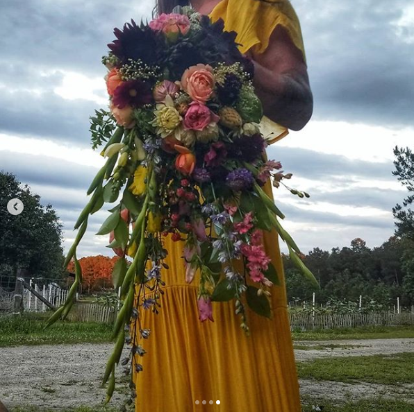 A woman in a yellow dress holds a messy cascade bouquet full of greenery with large leaves and pink, peach, purple, dark red, and small white flowers.