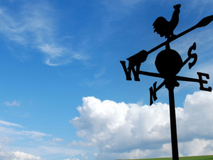 A silhouette of a weather vane with cardinal directions (N, S, E, W) and a rooster perched on top stands against a bright blue sky with scattered clouds. In the distance, a green field and a few bare trees are visible on the horizon.