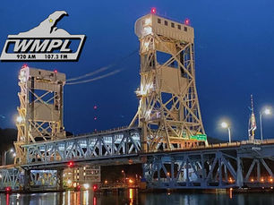 A nighttime photo of the Portage Lake Lift Bridge, brightly lit against a deep blue sky, is shown with the WMPL radio station logo in the top left corner. The logo includes the station frequencies 920 AM and 107.3 FM, along with an outline of Michigan’s Upper Peninsula.