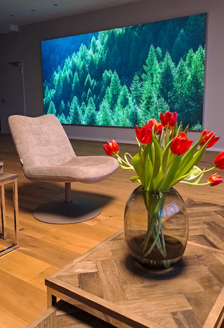 Rote Tulpen in Vase, daneben Sessel vor großem Bildschirm mit Waldmotiv.