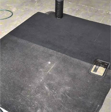 Tapete preto em piso de concreto, com poeira visível, um poste cilíndrico e uma alça metálica ao lado.