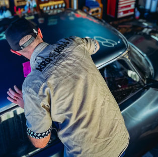 Homem em oficina polindo carro azul; camisa com KRAKOSS e GARAGE SOLUTIONS visíveis, clima de trabalho cuidadoso.