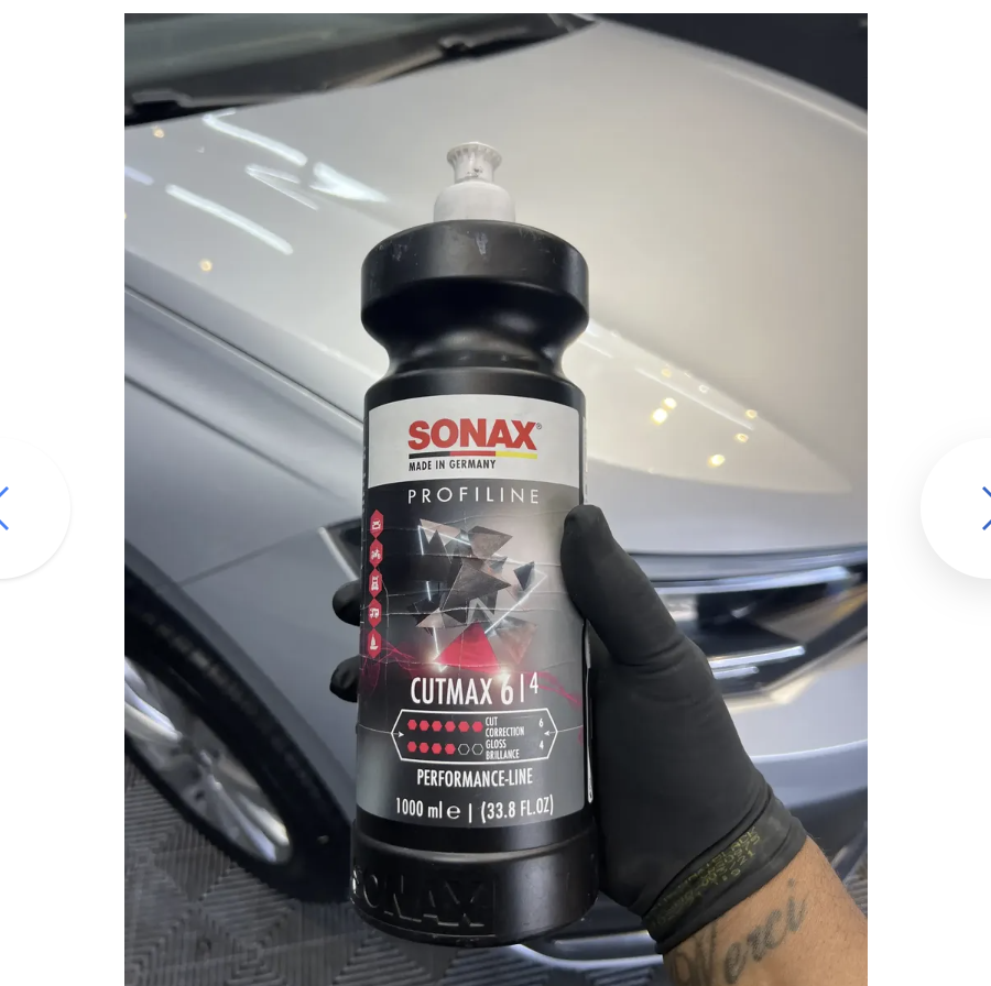 Mão com luva segura frasco SONAX CUTMAX 6|4 diante do capô de um carro branco em oficina.