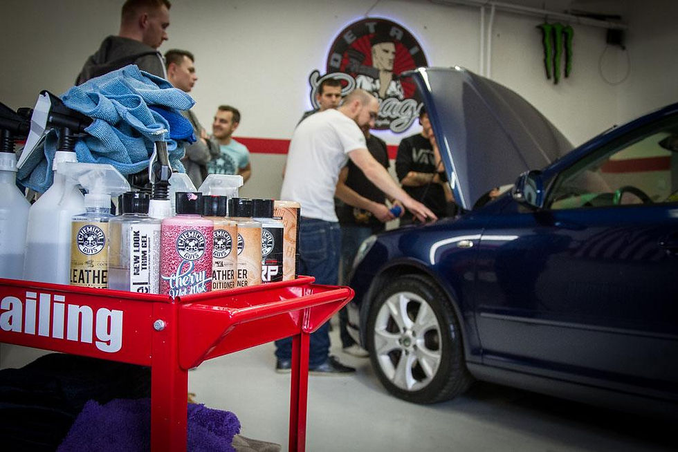Homens detalham um carro azul com capô aberto numa oficina; carrinho vermelho com produtos Chemical Guys em primeiro plano.