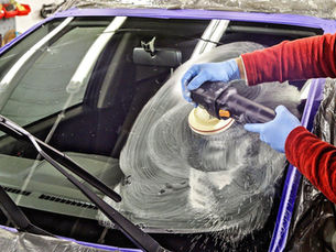 Mãos com luvas polindo o para-brisa de um carro roxo com politriz, em oficina, com espuma branca espirrando.