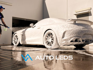 Carro esportivo coberto de espuma sendo lavado por profissional em lava-rápido, usando Snow Foam, com texto AUTO LEEDS na parte inferior.