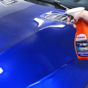 Mão pulveriza revestimento selante cerâmico SONAX no capô azul de carro, com brilho refletido e dutos de ar.
