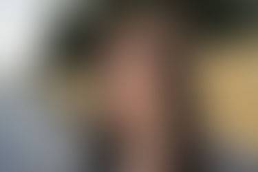 Mulher com expressão neutra em ambiente externo, carro azul ao fundo e árvores verdes. Blusa com padrão preto e branco, fundo de muro amarelo.