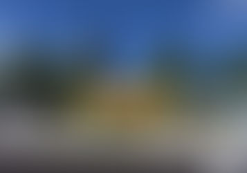 Praça colorida chamada "Praça dos Livros" com árvores ao fundo. Pessoas passeiam; um carro branco à direita. Céu azul claro.