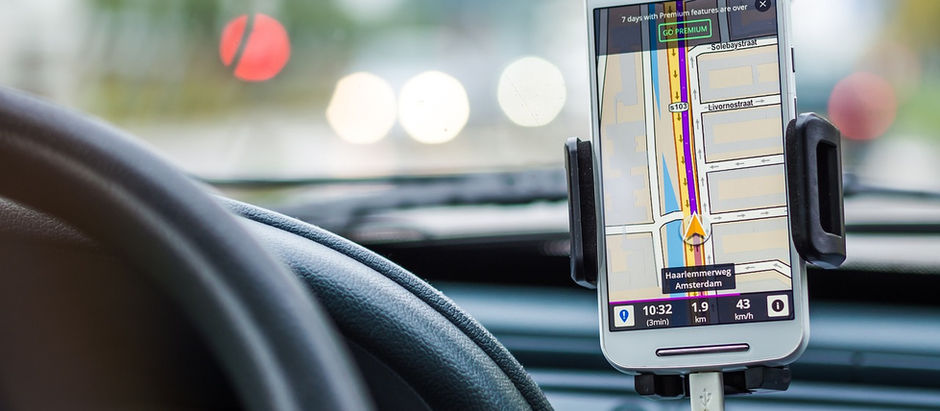 Smartphone com GPS mostrador em suporte no carro, exibindo trajeto em mapa. Volante à esquerda, fundo desfocado com luzes esmaecidas.