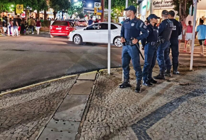 Policiais de uniforme azul monitoram movimentação noturna em rua com carros e pedestres. Loja e árvores iluminadas ao fundo.
