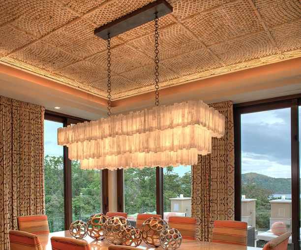 Three-tier rectangular selenite chandelier suspended above a luxury dining table in a coastal residence. Handcrafted from natural selenite crystal rods, the chandelier emits a warm ambient glow that complements the textured ceiling, custom furnishings, and panoramic ocean views.