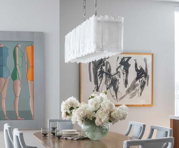 Rectangular selenite chandelier installed above a contemporary dining table, handcrafted from natural selenite crystals. The sculptural fixture emits a warm, diffused glow, creating an elegant focal point within the modern open-concept interior.