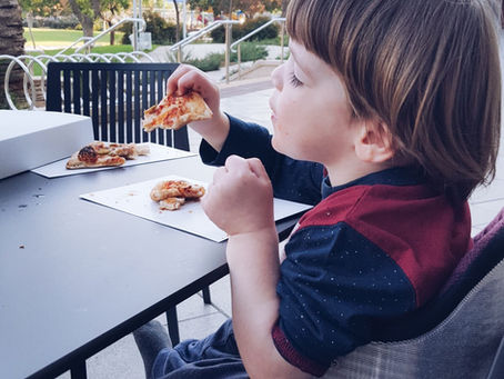 Child eating a slice of pizza at an outdoor table in a park on a sunny afternoon.