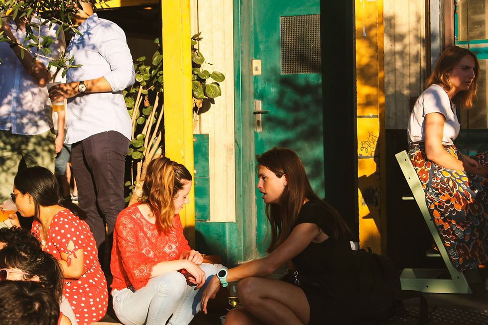 People enjoy an outdoor setting; two women chat on steps, one in red, one in black. A woman in a patterned skirt sits nearby. Sunlit and vibrant.