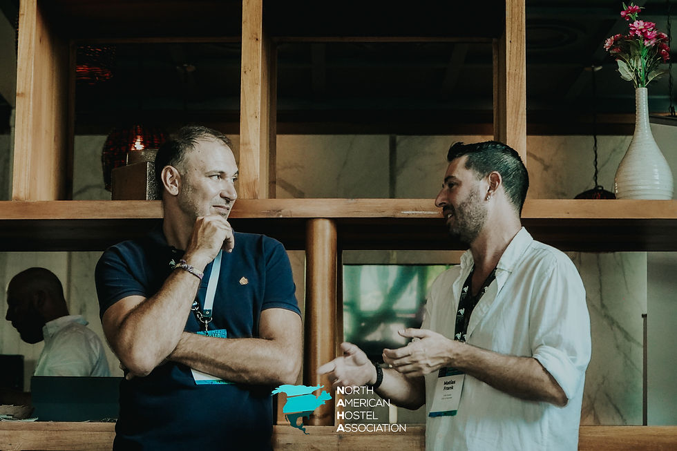 Two men engaged in conversation, one in a navy shirt, the other in a white shirt. Wooden shelves and a vase with pink flowers in background.