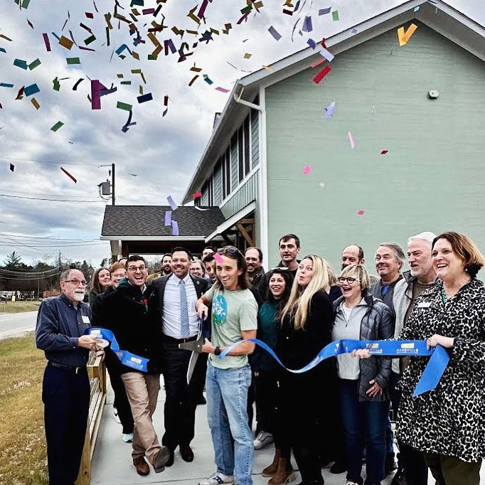 Group celebrating a ribbon-cutting ceremony outside a building. Colorful confetti falls as a person cuts a blue ribbon. Happy atmosphere.
