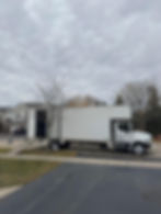 Moving truck parked in suburban driveway with a ramp, cloudy sky, bare trees, and houses in the background. A person is partially visible.