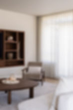 Lichte woonkamer met houten wandkast, fauteuil, ronde salontafel en kamerhoge gordijnen in een rustig interieur.