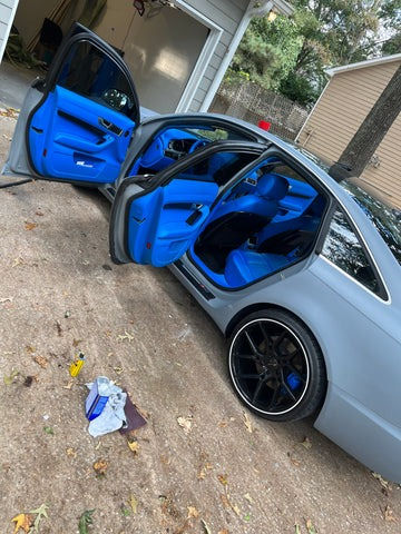 Car with open doors revealing bright blue interior, parked on a leaf-strewn driveway. Nearby cleaning supplies suggest detailing work.