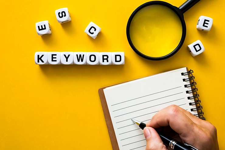 Hand writing on a notepad beside dice spelling "KEYWORD," a magnifying glass, and scattered letters on a yellow background.