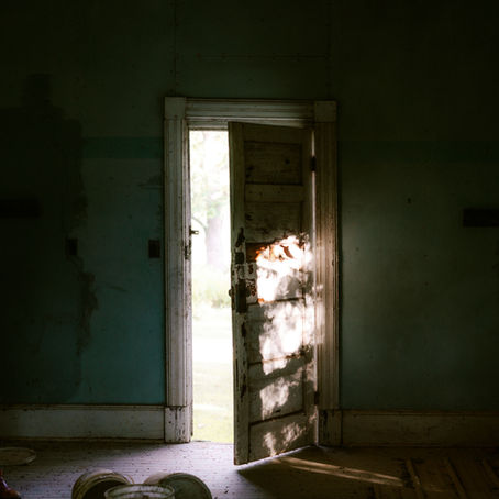 A partially open wooden door lets sunlight into a dim room, casting shadows on the floor representing the new beginnings of a solar eclipse in virgo. Two paint buckets rest on the worn, wooden floor.