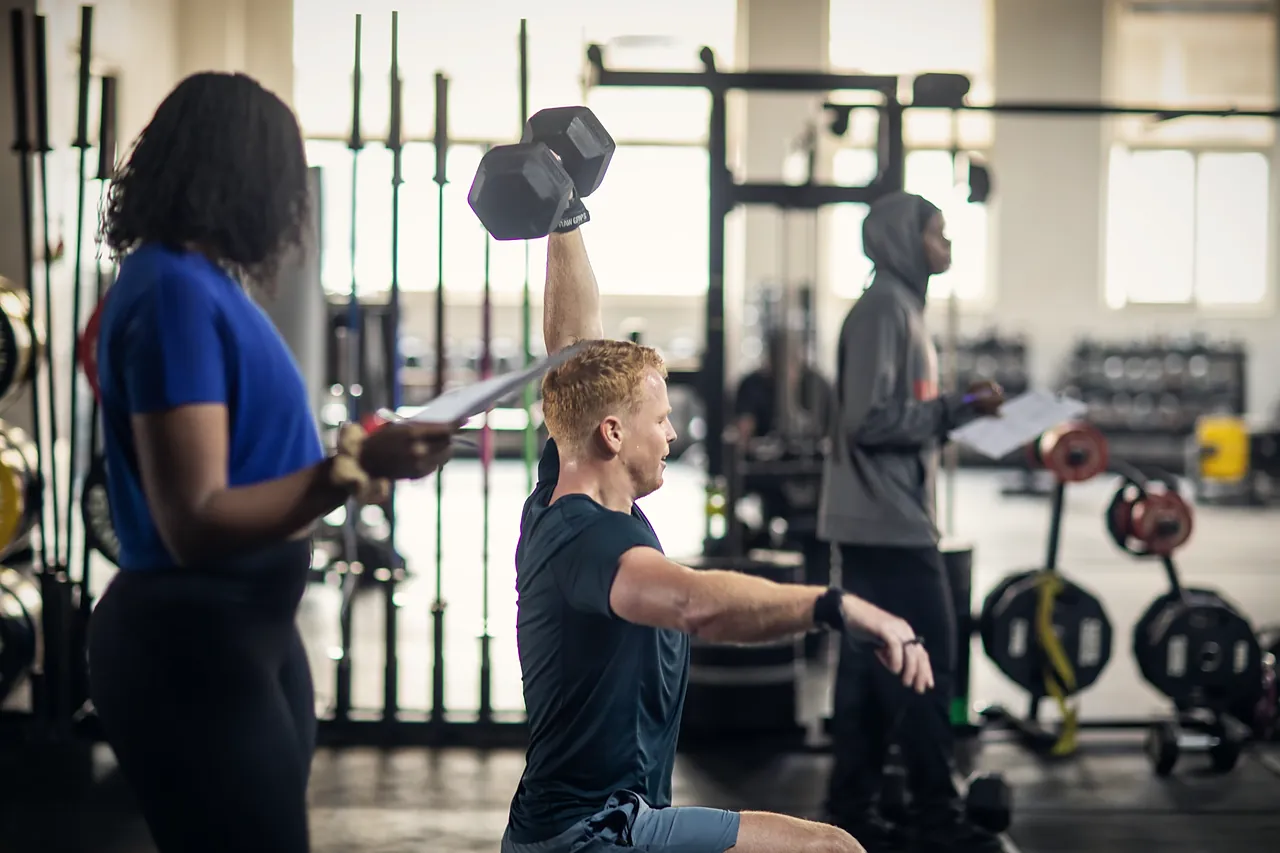 Coach recording results on a clipboard as a client presses a dumbbell overhead during an assessment