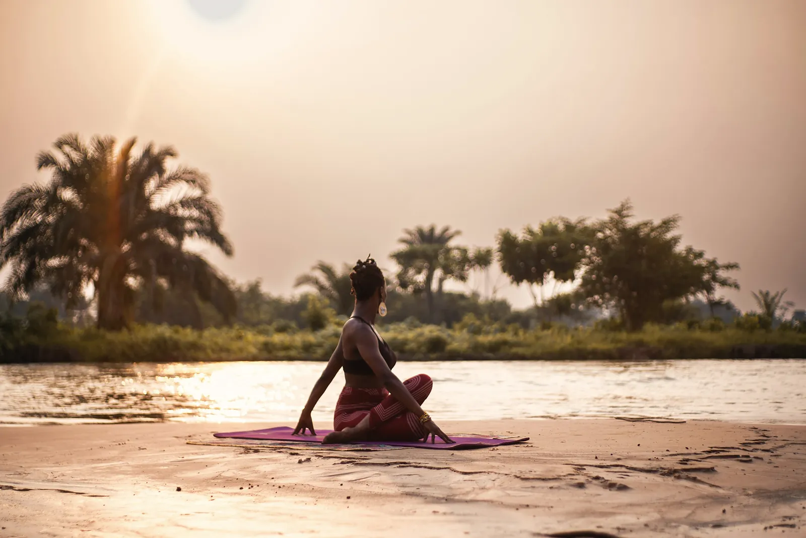 Woman holding a seated yoga twist on a mat by a river at sunrise