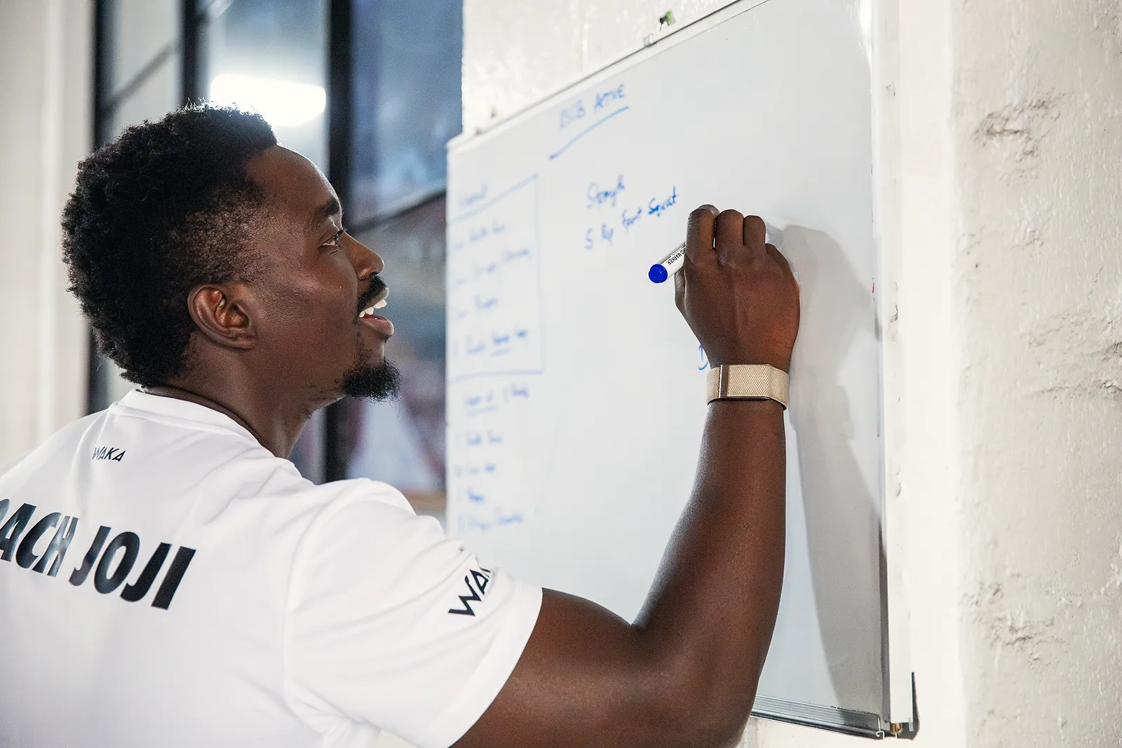 Coach Joji smiling as he writes a client’s strength session on a whiteboard