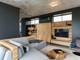 Modern living room with a gray sofa, wooden shelves, TV, and round wooden table on a beige rug. 