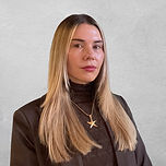 Portrait photo of a woman with brown-blonde chest-length hair, wearing a brown turtleneck jumper and a gold necklace