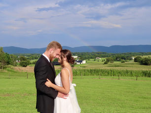 A bride and groom embrace in a sunlit field with rolling green hills behind them at Kateri Events and Weddings. A faint rainbow arcs across the sky above the distant Blue Ridge Mountains, adding a magical touch to the serene countryside estate setting.