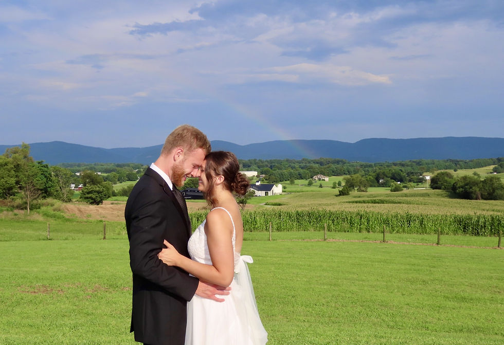 A bride and groom embrace in a sunlit field with rolling green hills behind them at Kateri Events and Weddings. A faint rainbow arcs across the sky above the distant Blue Ridge Mountains, adding a magical touch to the serene countryside estate setting.
