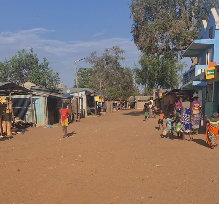 A dusty looking street with several small, man made aluminium buildings that are shops and restaurants. There are Malagasy families standing around or walking and doing their shopping