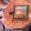 Thumbnail: A square Unakite pendant in a bronze-colored frame, showcasing a mix of earthy green and coral-pink tones with a reflective p