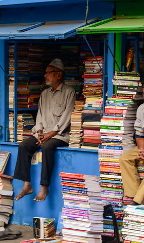 a cinematic street photography frame from college street, kolkata. it is one of the world's largest book market. in the frame a man sits in his book shop with religious cap on his head looking away.