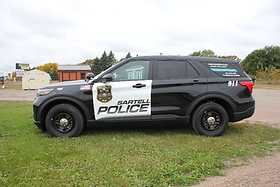 Side view of a Sartell Police SUV with a black and white vehicle wrap, featuring the police badge and lettering, parked on a grassy area.