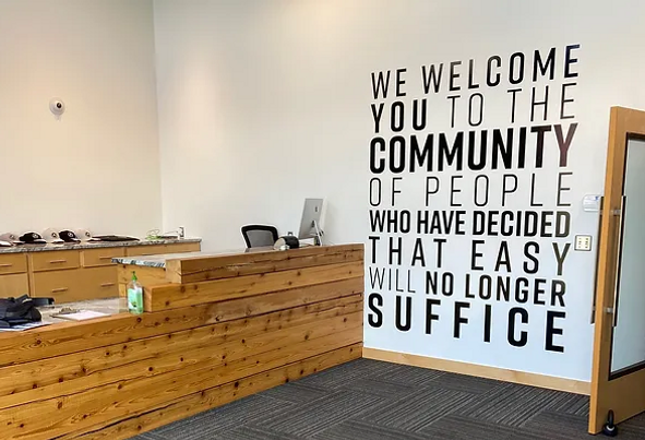 A motivational wall graphic in a modern office setting. The wall graphic reads, "We welcome you to the community of people who have decided that easy will no longer suffice," emphasizing a strong commitment to hard work and excellence. The office features a wooden reception desk and clean, professional decor.