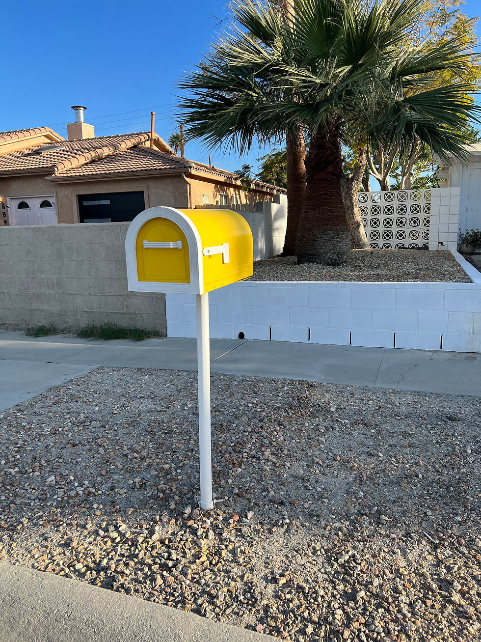 Yellow mailbox on a gravel patch by a sidewalk, under a palm tree. House with a red-tiled roof and a blue sky in the background.