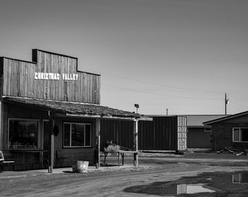 this is a picture of a old western style store, there's a small bench outfront, a wooden planter and this was taken in central oregon that was unused for awhile. there is a small pool of water out front and the image is in black and white. 