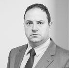 Advogado - homem branco de terno cinza e gravata, olhando seriamente para a câmera.