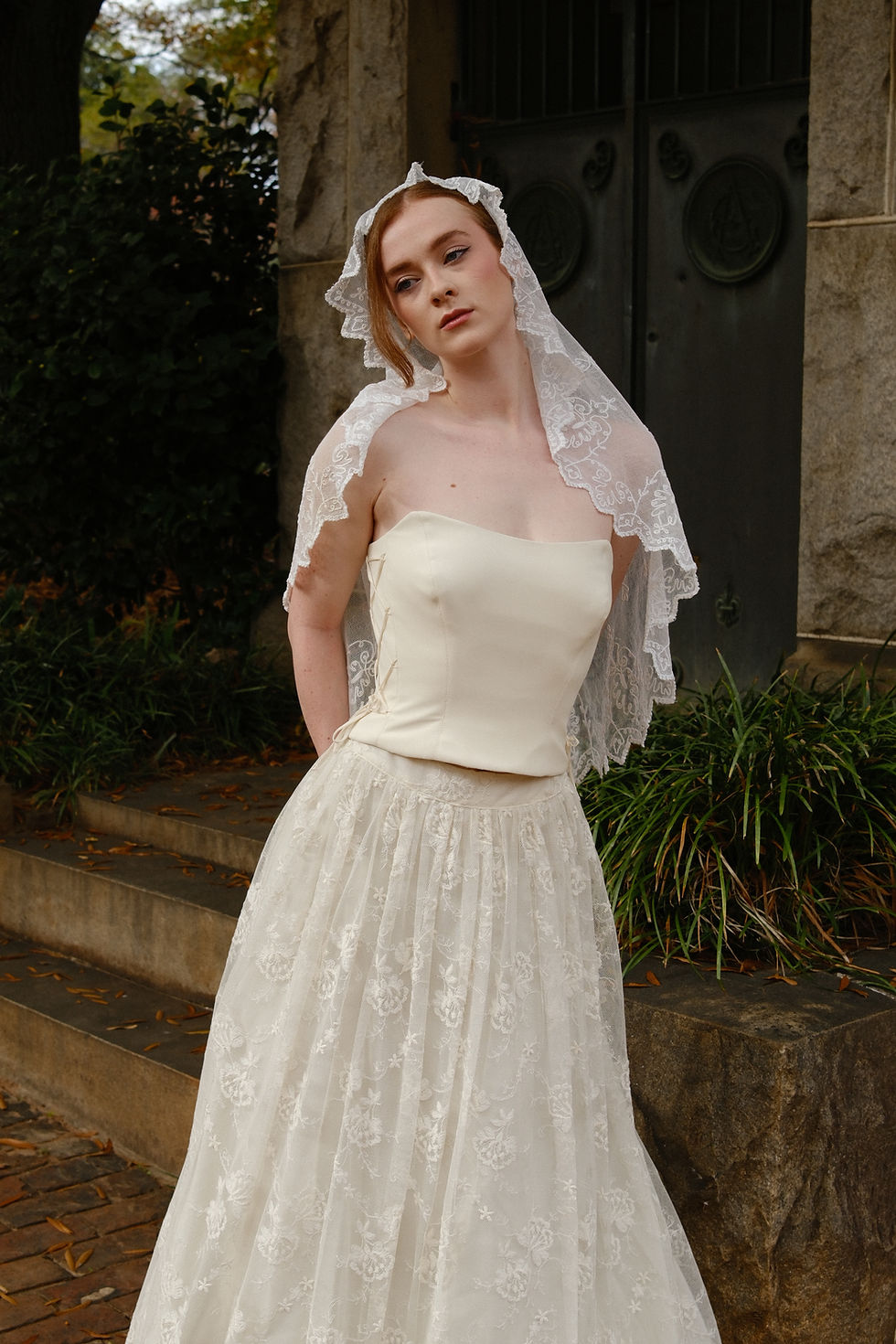 Bride wearing vintage wedding dress with corset and Italian lace skirt, standing near stone wall and black door