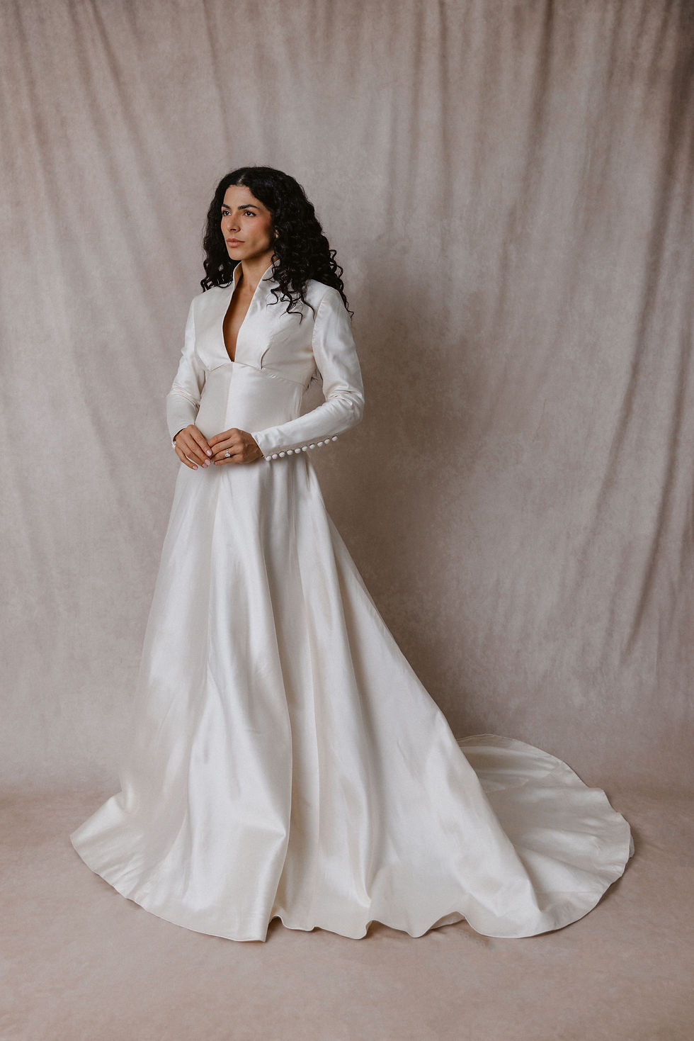 Model with dark curly hair wearing British vintage silk long sleeved wedding dress against beige backdrop