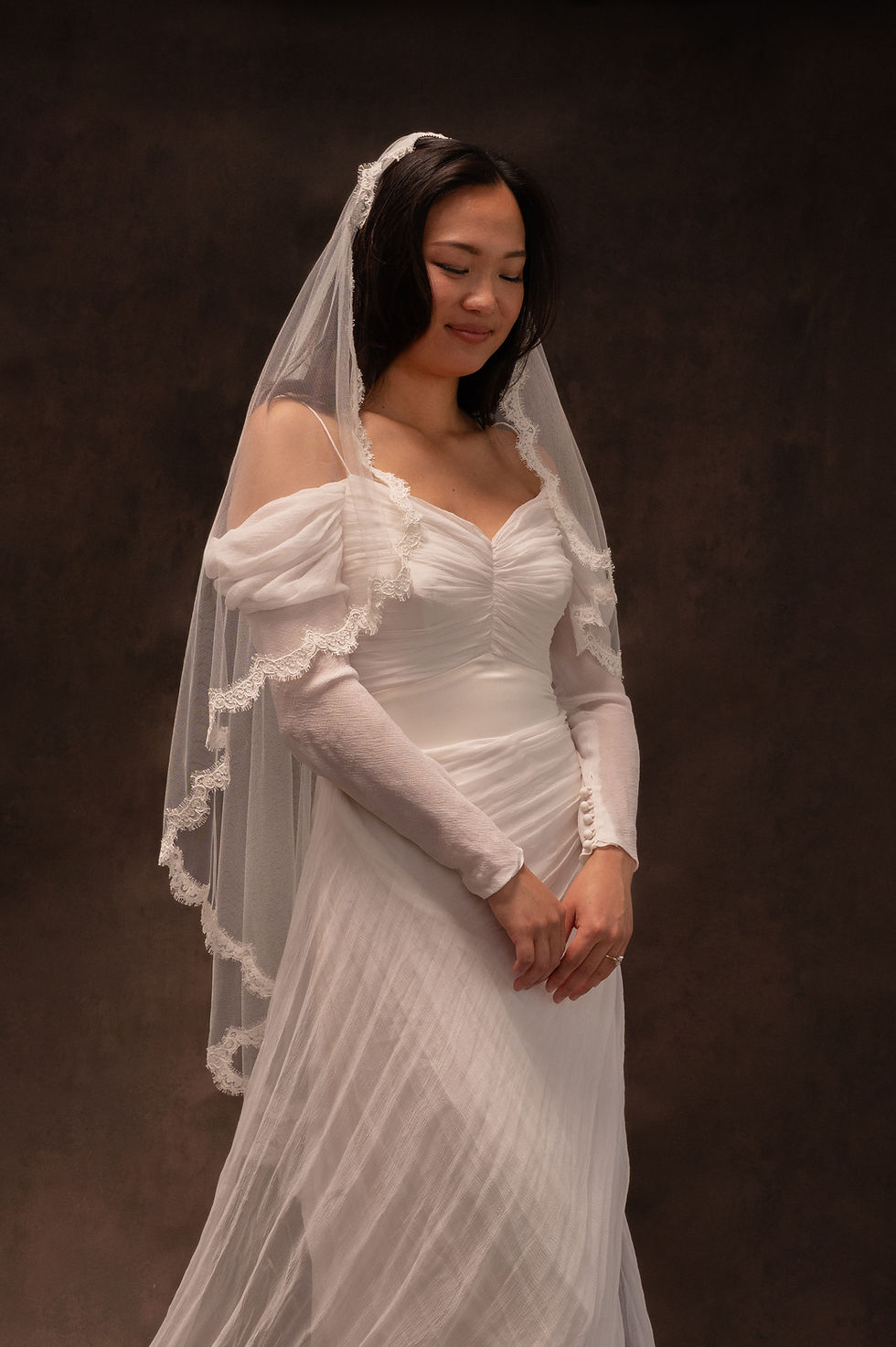 Thumbnail: Model wearing vintage Catherine Rayner wedding dress and lace-trimmed veil against brown backdrop