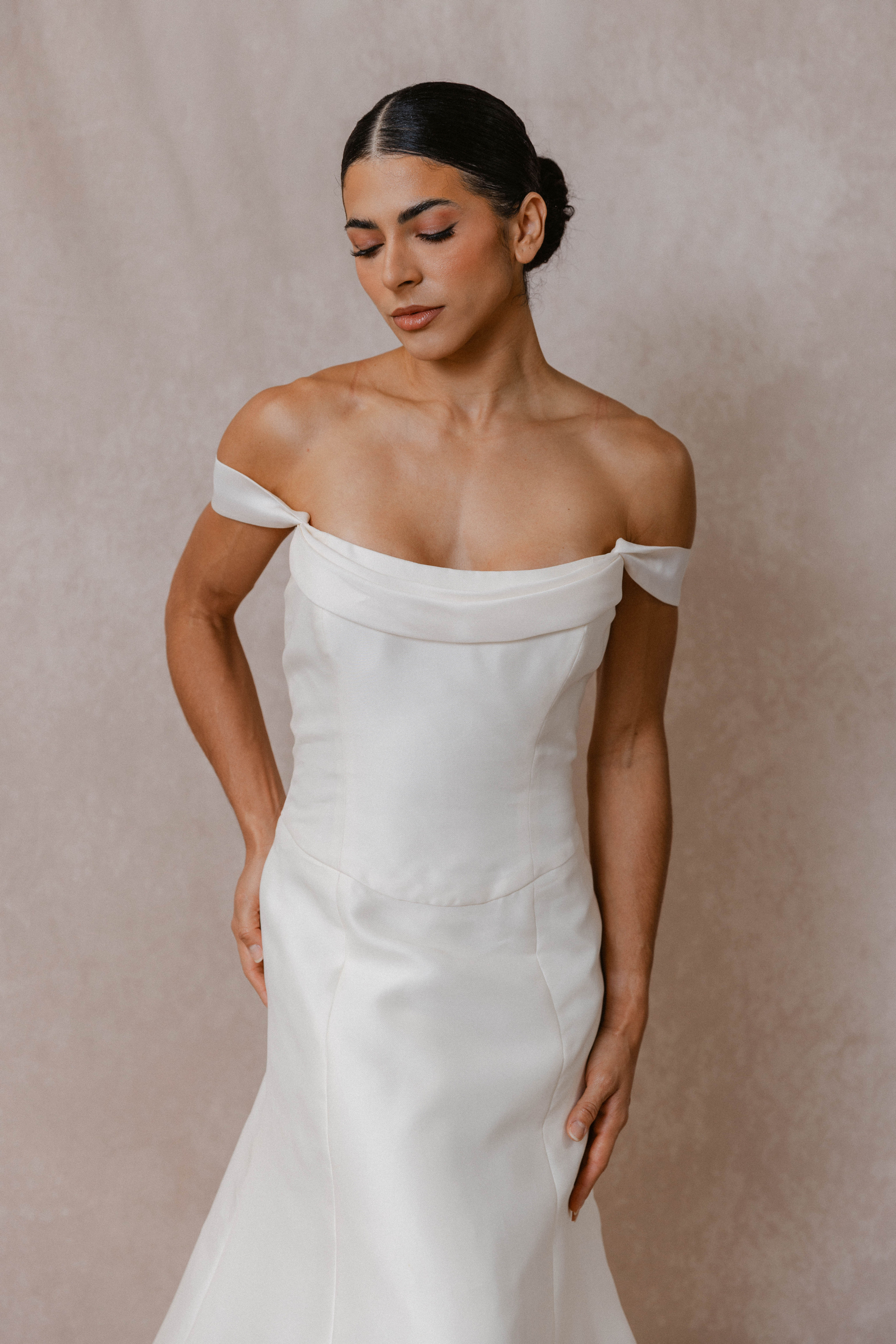 Dark haired woman standing against backdrop wearing vintage Vera Wang wedding dress with off shoulder strap and drop waist