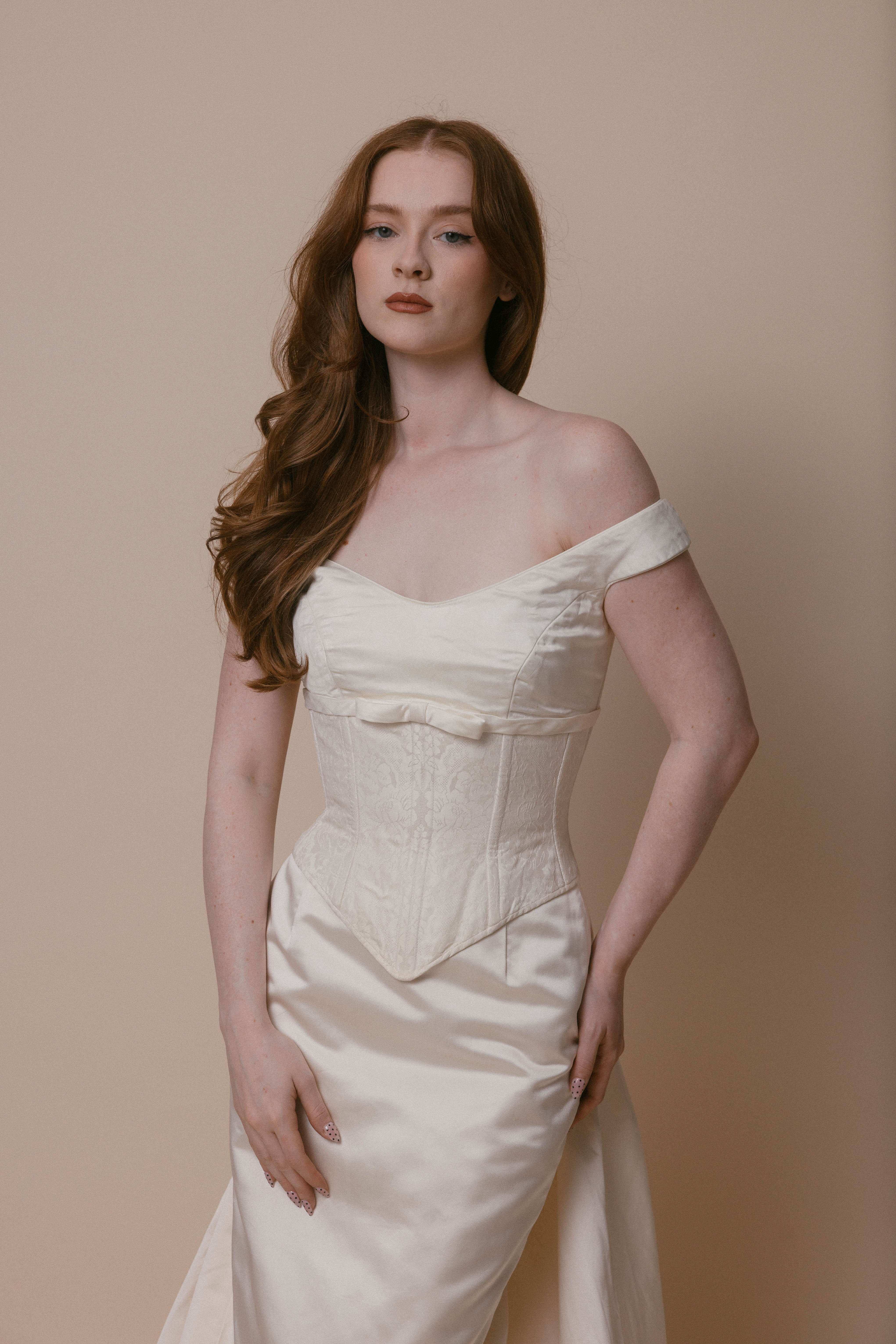 Red haired bride wearing vintage silk corset wedding dress and standing against an ivory backdrop